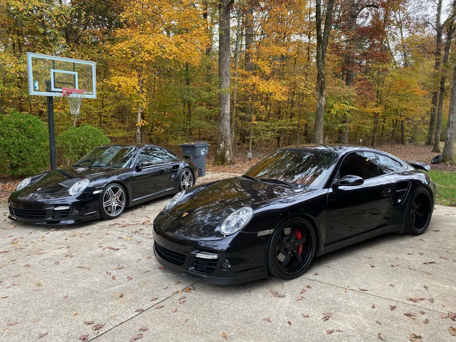 Two black Porsche 911 Turbo coupes detailed to a mirror gloss with fall foliage behind