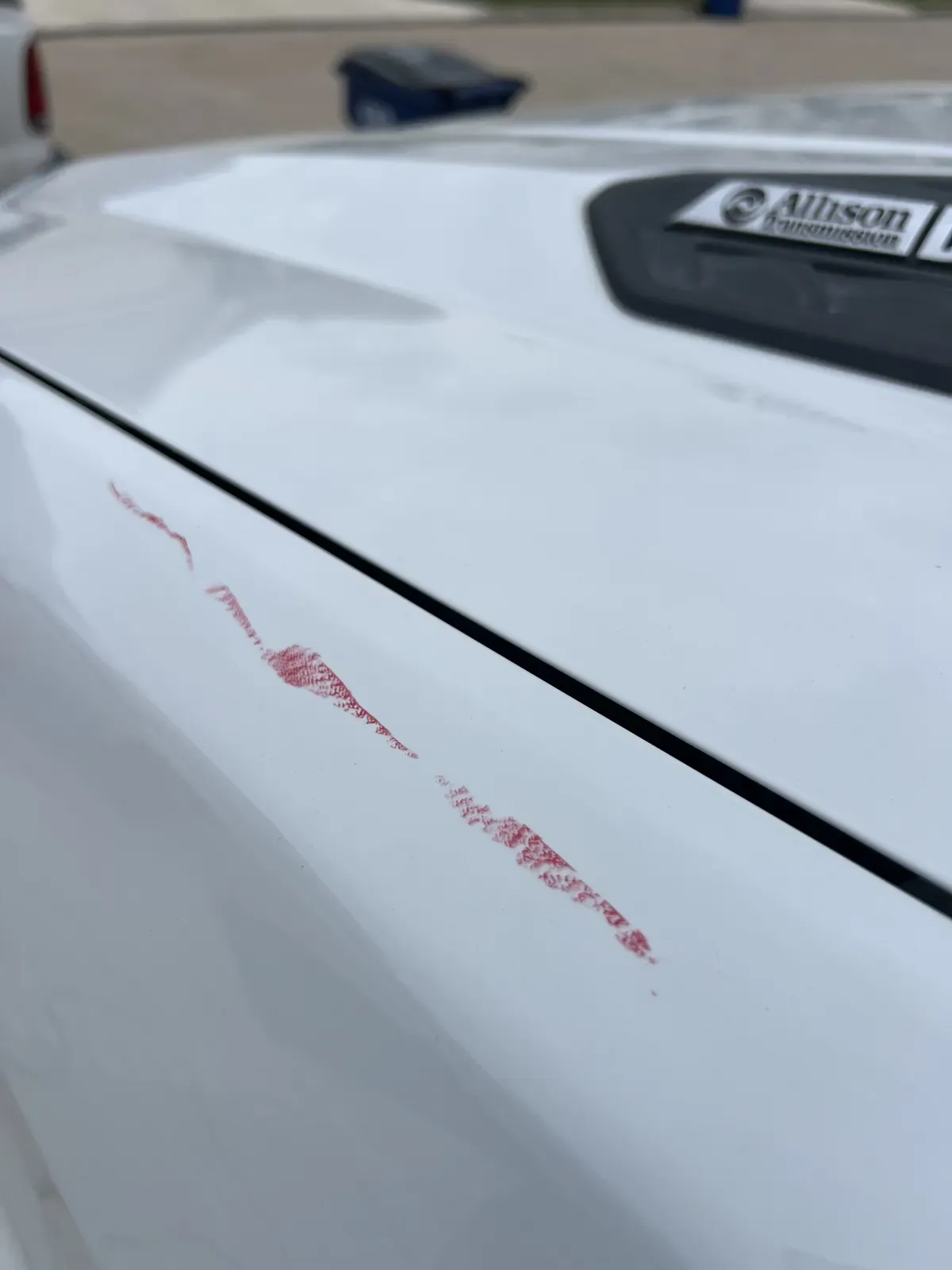 Red paint transfer streaked across the hood edge of a white Duramax pickup before correction